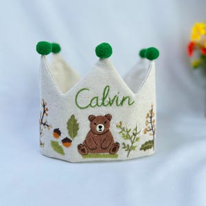 May include: A cream-colored fabric crown embroidered with the name "Calvin" in green script. The crown features a brown bear, acorns, and foliage embroidery. Green pom-poms adorn the tips of the crown.