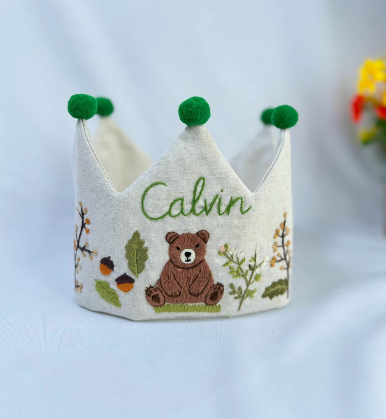 May include: A cream-colored fabric crown embroidered with the name "Calvin" in green script. The crown features a brown bear, acorns, and foliage embroidery. Green pom-poms adorn the tips of the crown.
