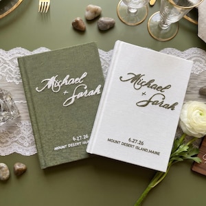 Pode incluir: Dois livros de visitas de capa dura com revestimento em tecido, um verde-oliva e outro branco. Ambos apresentam o texto bordado Michael + Sarah, a data 6.27.26 e a localiza&ccedil;&atilde;o Mount Desert Island, Maine. O livro verde tem texto branco, o livro branco tem texto verde-escuro.