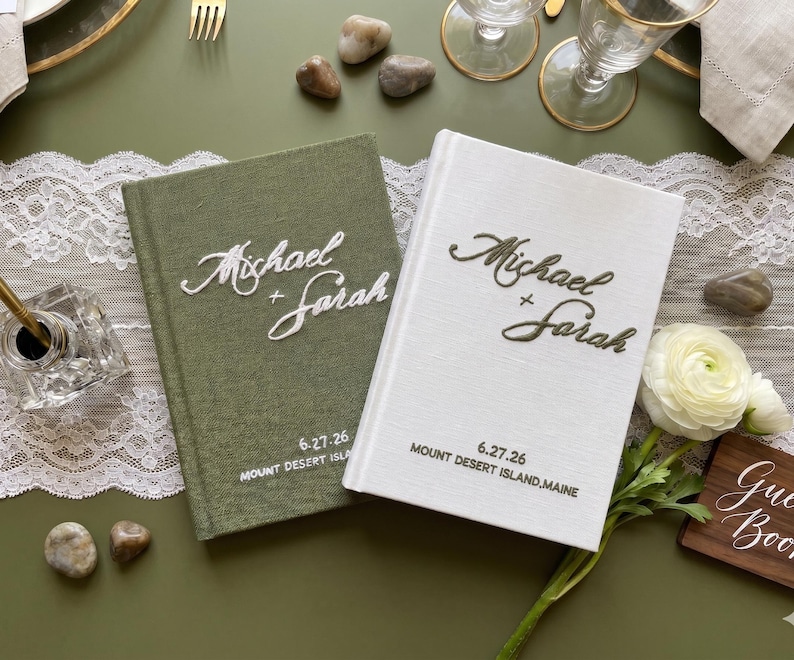 Pode incluir: Dois livros de visitas de capa dura com revestimento em tecido, um verde-oliva e outro branco. Ambos apresentam o texto bordado Michael + Sarah, a data 6.27.26 e a localiza&ccedil;&atilde;o Mount Desert Island, Maine. O livro verde tem texto branco, o livro branco tem texto verde-escuro.