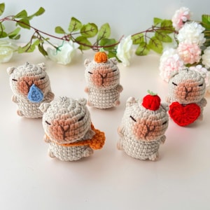 May include: Five crocheted stuffed animals in the shape of a llama with different accessories. The llama on the left has a blue teardrop, the llama in the middle has an orange scarf, the llama on the right has a red heart, the llama in the back left has an orange pumpkin, and the llama in the back right has a red strawberry.