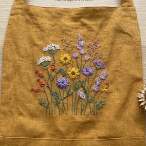 May include: A mustard yellow tote bag featuring a central hand-embroidered design of a wildflower bouquet. The embroidery includes white, yellow, orange, and purple flowers with green stems and leaves, stitched directly onto the fabric surface.