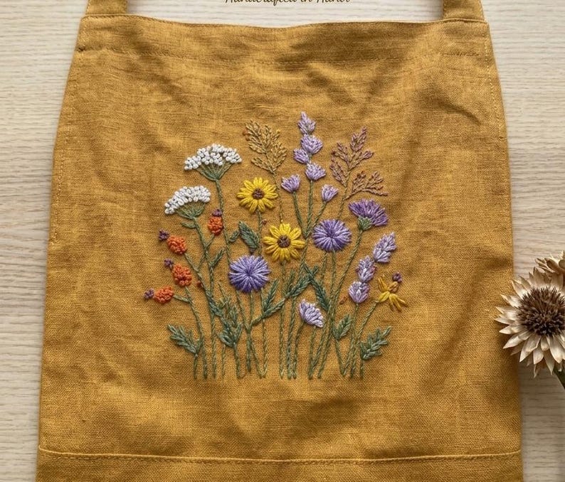 May include: A mustard yellow tote bag featuring a central hand-embroidered design of a wildflower bouquet. The embroidery includes white, yellow, orange, and purple flowers with green stems and leaves, stitched directly onto the fabric surface.