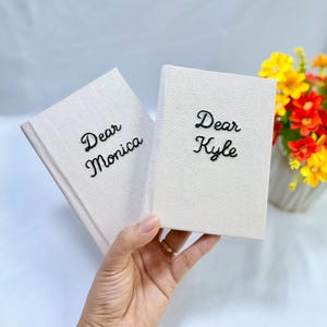 Puede incluir: Dos cuadernos rectangulares con cubiertas de tela color hueso. El cuaderno de la izquierda presenta el texto bordado en negro Dear Monica, mientras que el de la derecha presenta el texto bordado en negro Dear Kyle. Ambos artículos tienen un diseño sencillo.