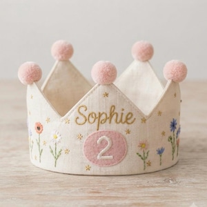 May include: A cream-colored fabric crown featuring embroidered wildflowers in shades of pink, white, and blue. The name Sophie is stitched in gold thread above a pink oval patch with the white number 2. Five pink pom-poms top the crown points.