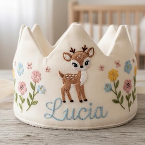 May include: Cream-colored crown with a deer and floral embroidery. The crown features a deer with brown spots and blue embroidered name "Lucia". The crown has a ribbon tie and is decorated with pink, blue, and yellow flowers.