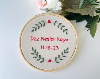 Può includere: Cerchio ricamato con una ghirlanda verde e bacche rosse. Il testo "Dez Nestor Kaye 11.18.23" è ricamato in rosso.