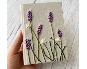 May include: A small, linen-covered notebook with embroidered purple lavender flowers and white daisies.