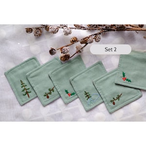 Può includere: Set di sei sottobicchieri in tessuto verde con disegni ricamati a tema invernale, tra cui un pino, un ramo di agrifoglio e un ramo di pino con una pigna. Il testo "Set 2" è visibile nell'immagine.