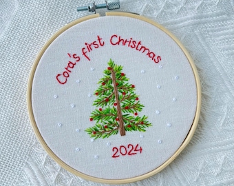 Può includere: Addobbo natalizio ricamato con un albero di Natale verde, bacche rosse e fiocchi di neve bianchi. Il testo "Cora's first Christmas 2024" è ricamato in filo rosso.