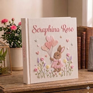 Puede incluir: Un libro color crema con el nombre "Seraphina Rose" bordado en rosa. La portada presenta un conejito bordado sosteniendo globos rosas en forma de coraz&oacute;n, rodeado de flores y estrellas. La columna vertebral tiene el a&ntilde;o "2026".