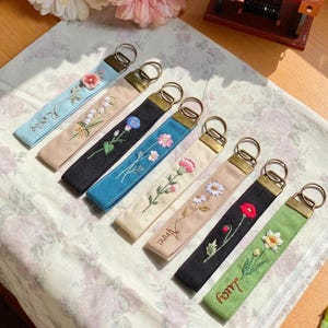 Pode incluir: Uma coleção de chaveiros bordados em várias cores, incluindo azul, bege, preto e verde. Cada chaveiro apresenta um design floral diferente e um anel de metal dourado. Alguns chaveiros têm nomes bordados.