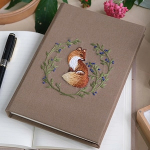 May include: A brown linen journal featuring an embroidered fox and blue berries. The fox is sitting within a wreath of green leaves and blue berries.