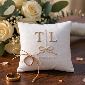May include: A square, off-white ring pillow with embroidered initials "T|L", a bow, and the date "12.05.2027". A gold wedding ring is tied to the pillow with twine. A second gold ring rests on the wooden surface. A bouquet of white roses is in the background.