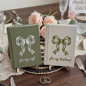 Pode incluir: Dois livros de capa dura com revestimento em tecido. O livro verde-oliva apresenta um la&ccedil;o branco bordado e o texto "for my wife". O livro de cor creme apresenta um la&ccedil;o verde-oliva bordado e o texto "for my husband". Ambos os livros est&atilde;o em p&eacute;.