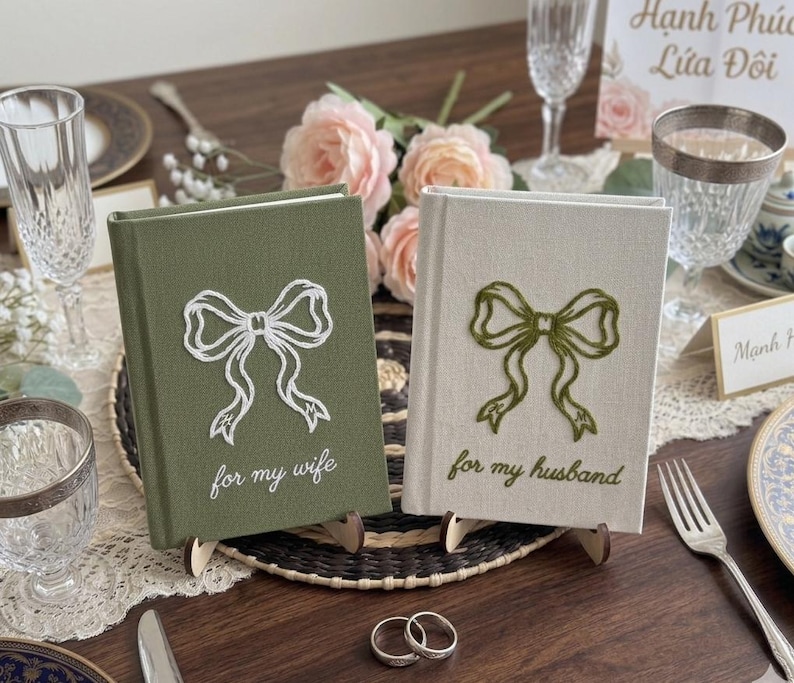 Pode incluir: Dois livros de capa dura com revestimento em tecido. O livro verde-oliva apresenta um la&ccedil;o branco bordado e o texto "for my wife". O livro de cor creme apresenta um la&ccedil;o verde-oliva bordado e o texto "for my husband". Ambos os livros est&atilde;o em p&eacute;.