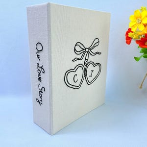 May include: A cream-coloured photo album with the words "Our Love Story" in script on the spine. The front features a black embroidered design of two connected hearts with the letters "C" and "I" inside, and a bow.