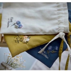 May include: Three fabric drawstring bags with embroidered floral designs. The top bag is white with a blue and purple flower and the text "Petite Flora". The middle bag is yellow with a yellow, orange, and white flower. The bottom bag is blue with a purple flower and the text "Via Lactea".