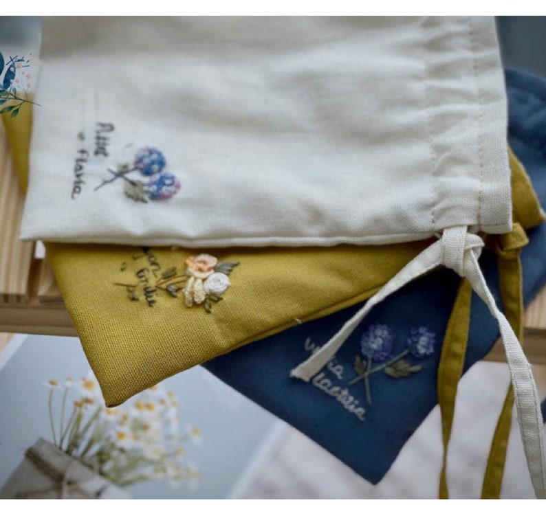 May include: Three fabric drawstring bags with embroidered floral designs. The top bag is white with a blue and purple flower and the text "Petite Flora". The middle bag is yellow with a yellow, orange, and white flower. The bottom bag is blue with a purple flower and the text "Via Lactea".