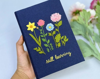 May include: A navy blue journal with embroidered flowers in yellow, pink, and blue. The words "still learning" are embroidered in gold thread. The journal is held in a hand.