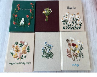 May include: Six embroidered notebooks with different floral designs. The covers are beige, green, and brown. The titles on the covers are "The Garden", "Hazel Bee", "my journey to baby sigart", "indigo", and two others that are not visible.