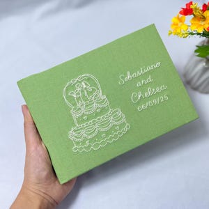 Op de afbeelding: Een lichtgroen, rechthoekig trouwgastenboek met witte geborduurde details. Het ontwerp toont een bruidstaart en de namen "Sebastiano and Chelsea" met de datum "06/09/25". Het boek wordt in een hand gehouden.