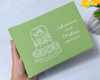 May include: A light green, rectangular wedding guest book with white embroidered details. The design features a wedding cake and the names "Sebastiano and Chelsea" with the date "06/09/25". The book is held in a hand.