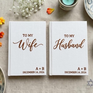 Pode incluir: Dois livros de capa dura brancos com texto castanho. O livro da esquerda diz TO MY Wife A + B DECEMBER 14, 2026. O livro da direita diz TO MY Husband A + B DECEMBER 14, 2026. As capas t&ecirc;m um acabamento em tecido texturizado.