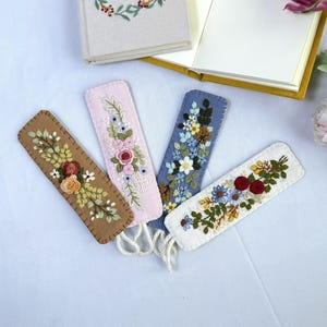 K&ouml;nnte beinhalten: Vier bestickte Filz-Lesezeichen in Braun, Rosa, Blau und Wei&szlig;, jeweils mit floralen Designs. Die Lesezeichen haben cremefarbene Quasten und liegen in der N&auml;he eines offenen und eines geschlossenen Buches.