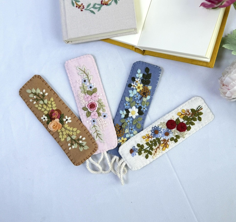 K&ouml;nnte beinhalten: Vier bestickte Filz-Lesezeichen in Braun, Rosa, Blau und Wei&szlig;, jeweils mit floralen Designs. Die Lesezeichen haben cremefarbene Quasten und liegen in der N&auml;he eines offenen und eines geschlossenen Buches.
