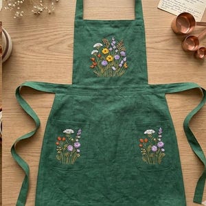 May include: A dark green apron featuring embroidered wildflower bouquets on the chest and two front pockets. The embroidery includes yellow, white, orange, and purple flowers with green stems. The apron has long ties and a neck strap.