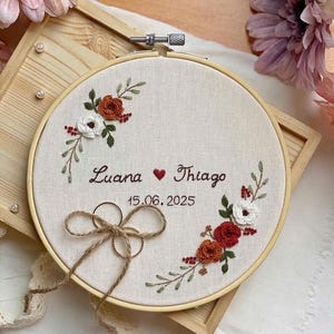 Op de afbeelding: Geborduurde borduurring met de namen "Luana" en "Thiago", een hartsymbool en de datum "15.06.2025". Het ontwerp toont bloemenborduurwerk in rood, oranje en wit, met een touwstrik en twee ringen. De ring zit in een houten frame.