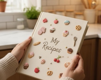 Puede incluir: Un libro de recetas color crema titulado "My Recipes" con decoraciones en relieve con temática de comida. La portada presenta cupcakes, galletas y frutas en miniatura. Un batidor y pan también están incluidos en el diseño. El libro está abierto, mostrando las páginas interiores.