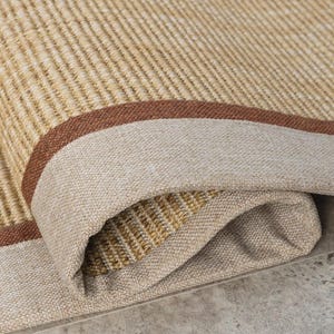 May include: A woven area rug featuring a textured tan and beige surface with a tight, linear pattern. The rug has a wide, solid beige fabric border framed by a thin, dark brown stripe, with one corner folded over to reveal the underside.