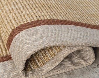 Puede incluir: Una alfombra tejida con una superficie texturizada en tonos bronceado y beige con un patrón lineal apretado. La alfombra tiene un borde ancho de tela beige sólida enmarcado por una fina raya marrón oscuro, con una esquina doblada.