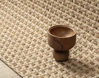 Puede incluir: Un pequeño cuenco de madera redondeado con un acabado liso y pulido y una base esférica gruesa, apoyado sobre una alfombra de color tostado tejida y texturizada con un patrón de rejilla repetitivo.