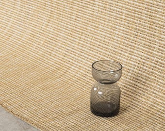 May include: A small, hourglass-shaped glass vase with a dark gray, translucent tint. The vase features a rounded base and a wider, open top, resting on a textured, light tan woven rug with a visible horizontal ribbed pattern.
