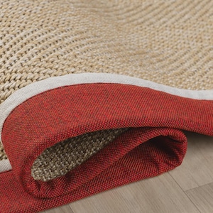 May include: Close-up of a rolled-up area rug. The rug features a woven beige center, a red border, and a neutral-colored trim. The rug is on a light-colored wooden floor.