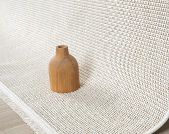 May include: A light beige woven area rug with a textured grid pattern and a short fringe edge, featuring a small, smooth, light brown wooden vase placed on its surface. The rug is partially rolled up, revealing its structure.