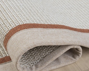Puede incluir: Una alfombra tejida con un centro texturizado de color crema y un borde ancho de color beige claro. Una banda estrecha y distintiva de tela naranja quemado separa el centro crema del borde beige, creando un borde doblado y superpuesto.