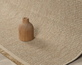May include: A beige woven area rug with a textured grid pattern and a short fringe edge, featuring a small, smooth, light brown wooden vase placed on its surface. The rug has a consistent neutral tone and a dense, uniform weave structure.