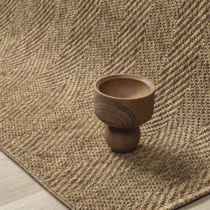 May include: A small, brown, turned wooden bowl with a rounded, pedestal-style base. The bowl features a smooth, polished finish and a deep, circular interior. It is positioned on a textured, tan-colored rug with a prominent herringbone pattern.