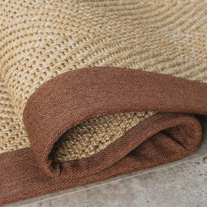 Puede incluir: Primer plano de una alfombra enrollada y tejida con un centro beige natural y un borde marrón oscuro. La textura de la alfombra es visible, mostrando el patrón tejido y los colores contrastantes.