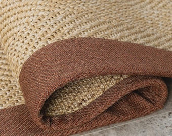 Puede incluir: Una alfombra tejida que presenta una superficie de fibra natural de color tostado con textura y un patrón de espiga distintivo. La alfombra está rematada con un borde de tela ancho y sólido de color marrón oscuro.