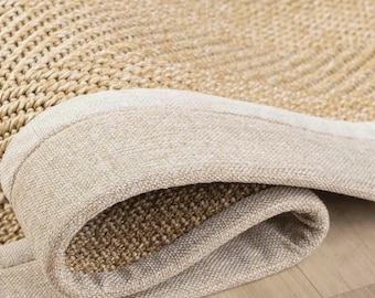 May include: A woven area rug featuring a textured, tan-colored herringbone pattern. The rug has a thick, light beige fabric border that wraps around the edge, creating a folded corner that reveals the dense, natural fiber construction underneath.