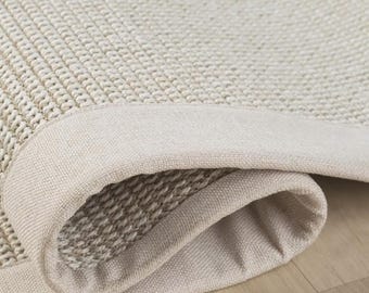 Puede incluir: Una alfombra beige con un tejido denso y texturizado y un borde de tela lisa de color sólido. La alfombra está parcialmente doblada, revelando la parte inferior y el patrón tejido apretado y consistente del material.