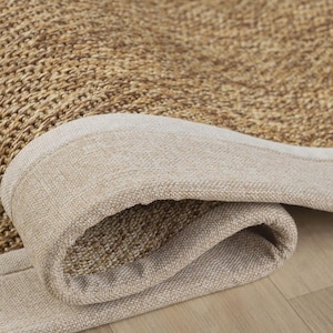 Puede incluir: Una alfombra tejida con una superficie de fibras naturales de color tostado con un patrón trenzado apretado. La alfombra está rematada con un borde de tela ancho, liso y de color beige claro que envuelve el borde, mostrando un detalle de esquina doblada.
