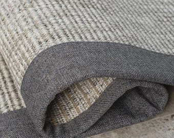 Puede incluir: Una alfombra tejida con una superficie texturizada de color beige claro y crema con un borde de tela gris carbón oscuro. La alfombra está parcialmente doblada, revelando el contraste entre el centro tejido claro y el borde oscuro.