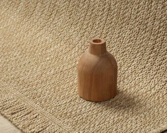 May include: A tan woven rug with a textured surface and a fringed edge, featuring a small, smooth, light brown wooden vase with a narrow neck and a rounded body placed directly on top of the rug's surface.