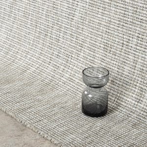 May include: A small, hourglass-shaped glass vase with a dark gray gradient finish, positioned on a textured, woven rug featuring a neutral beige and white horizontal striped pattern that extends across the surface in a continuous, repeating weave.